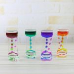 Quatro ampulhetas de óleo líquido com cores diferentes: rosa, verde, azul e laranja, exibidas numa superfície de madeira clara contra um fundo de parede branca.
