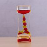 Ampulheta com óleo líquido vermelho e bolinhas amarelas a fluir de cima para baixo, visível através de um vidro curvo transparente.
