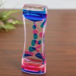 Ampulheta de óleo líquido com cores azul, rosa e transparente, mostrando o movimento do óleo dentro do vidro curvado sobre uma superfície de madeira.