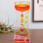 Ampulheta de óleo líquido com cores rosa, amarelo e laranja, mostrando o fluxo de óleo de cima para baixo em um recipiente de vidro transparente.