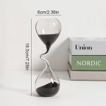 Ampulheta com forma irregular, com areia preta, altura de 18,5cm e largura de 6cm, colocada ao lado de caixas de papel com marcas Union e Nordic.