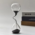 Ampulheta com areia preta, estrutura de vidro com forma irregular, fundo branco, livros ao lado, destaque para o design único da ampulheta.
