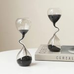 Ampulhetas de vidro com areia preta, uma com forma tradicional e outra com forma ondulada, colocadas sobre um livro de ciência.