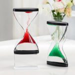 Duas ampulhetas mágicas invertidas com areia colorida, uma com areia vermelha e outra com areia verde, em diferentes ângulos, sobre uma mesa branca com flores ao fundo.