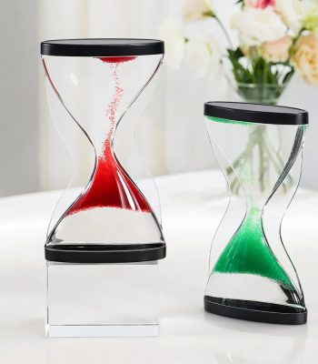 Duas ampulhetas mágicas invertidas com areia colorida, uma com areia vermelha e outra com areia verde, em diferentes ângulos, sobre uma mesa branca com flores ao fundo.