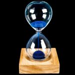 Ampulheta magnética com areia azul em vidro transparente, apoiada numa base de madeira, com reflexos de luz na superfície de vidro.