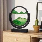 Lâmpada decorativa com painel de paisagem montanhosa verde, estrutura circular preta, colocada numa mesa de madeira com uma planta de cacto ao lado.