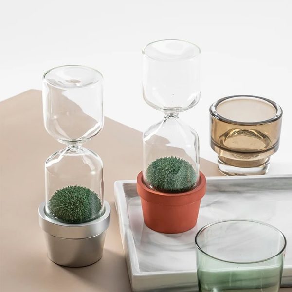 Vasos de vidro com plantas verdes em diferentes suportes, incluindo um vaso de barro e um recipiente de metal, dispostos sobre uma bandeja de mármore e uma superfície de cor bege.