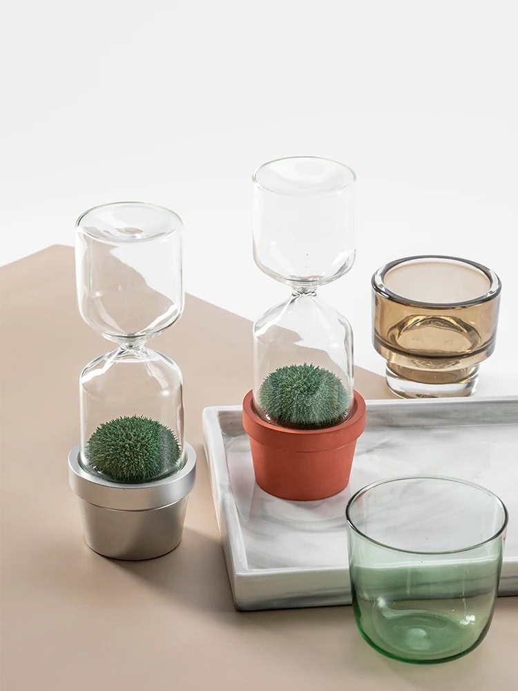 Vasos de vidro com plantas verdes em diferentes suportes, incluindo um vaso de barro e um recipiente de metal, dispostos sobre uma bandeja de mármore e uma superfície de cor bege.