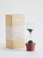 Ampulheta com vaso de flores, areia negra a fluir, embalagem de papel kraft ao lado, fundo branco.
