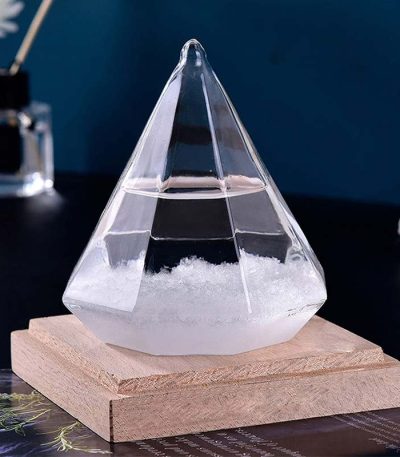 Cristais transparentes em forma de pirâmide com base de madeira, usado como barómetro de ciência, com fundo escuro e objetos de decoração ao fundo.