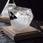 Cristal transparente com líquido, montado numa base de madeira, usado para demonstrações científicas de pressão atmosférica.