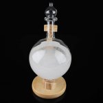 Frasco de vidro com líquido branco e esfera de cristal, base de madeira, usado para demonstrações científicas.