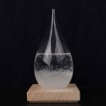 Cristal de gota de água com líquido transparente, forma alongada, colocado sobre base de madeira escura, fundo preto.