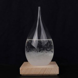 Cristal de gota de água com líquido transparente, forma alongada, colocado sobre base de madeira escura, fundo preto.