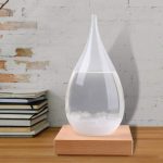 Barómetro de cristais em forma de gota de água, com líquido transparente e cristais brancos na base, colocado sobre uma plataforma de madeira.