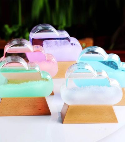 Conjunto de cinco nuvens de cristal de cores diferentes, cada uma colocada sobre uma base de madeira, com fundo de plantas verdes.