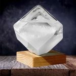 Cristal de sal de Rocha transparente com forma de cubo, colocado sobre uma base de madeira, com fundo escuro e iluminação que realça a sua transparência e detalhes.