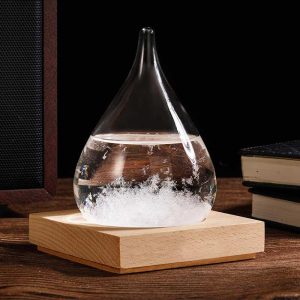 Gota de vidro com cristais de tempestade no interior, sobre base de madeira, com livros ao fundo.
