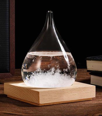 Gota de vidro com cristais de tempestade no interior, sobre base de madeira, com livros ao fundo.