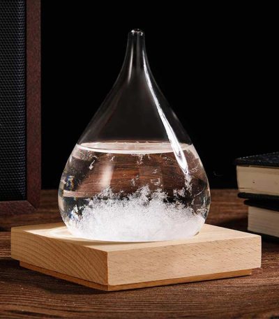 Gota de vidro com cristais de tempestade no interior, sobre base de madeira, com livros ao fundo.