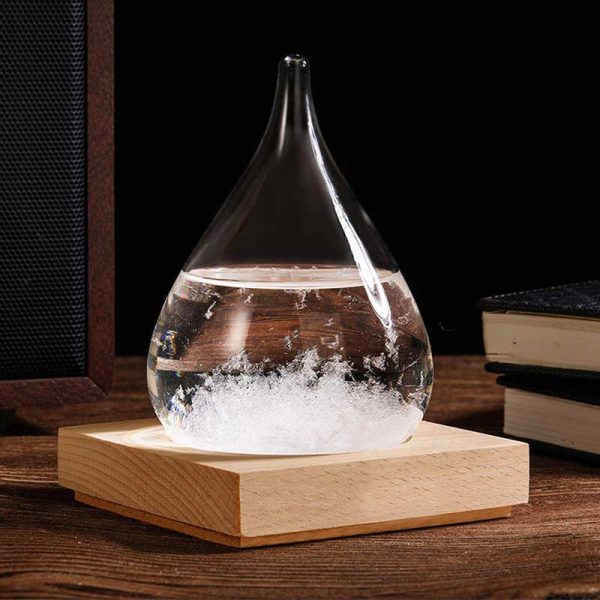 Gota de vidro com cristais de tempestade no interior, sobre base de madeira, com livros ao fundo.