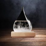 Cristal de forma gota com efeito de tempestade, parcialmente cheio de líquido, colocado numa base de madeira escura.