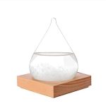 Gota de vidro com água e cristais de sal, suspensa por fio fino, sobre base de madeira, com fundo branco.