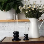 Dois tubos de vidro de diferentes tamanhos com bases de madeira escura, colocados numa mesa de madeira, com uma jarra branca de flores ao fundo.