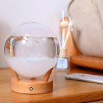 Barómetro decorativo com esfera de vidro transparente, base de madeira, e líquido visível, colocado numa mesa de madeira com objetos de decoração ao fundo.