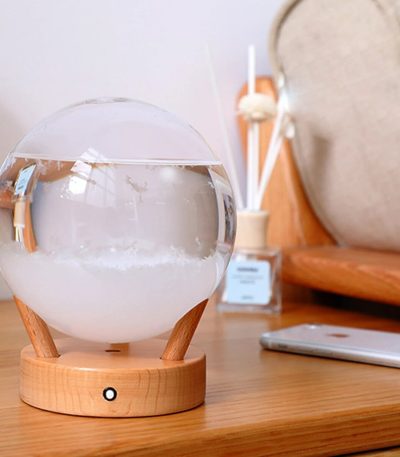 Barómetro decorativo com esfera de vidro transparente, base de madeira, e líquido visível, colocado numa mesa de madeira com objetos de decoração ao fundo.