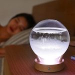 Barómetro decorativo em forma de bola de cristal com base de madeira, iluminado por luz LED, com uma mulher a dormir ao fundo.
