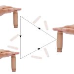 Duas mãos segurando bastões de madeira Mokuru, com várias peças ao redor, demonstrando o movimento de equilíbrio e manipulação do brinquedo.