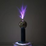 Bobina de Tesla com faíscas violetas a emergir do topo, refletidas na esfera metálica, com base iluminada por luzes azuis.