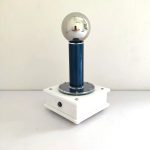 Bobina de Tesla com esfera metálica refletindo o ambiente, montada numa base branca com detalhes em azul e metal.