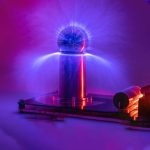 Bobina de Tesla Mini a gerar uma descarga de plasma azul e roxa, com faíscas visíveis na ponta, sobre uma base escura com iluminação colorida.