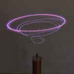 Efeito de descargas elétricas em forma de anéis concêntricos de cor violeta, gerados por uma bobina de Tesla miniatura, com foco na descarga e no dispositivo na base.
