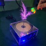 Bobina de Tesla musical com plasma colorido e luzes LED, mostrando fios de plasma brilhantes e uma mão segurando LEDs coloridos ao lado do dispositivo.