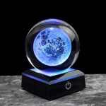 Lâmpada de cristal com efeito lunar azul, base preta com botão de ligar, iluminação suave e detalhes da superfície lunar visíveis.