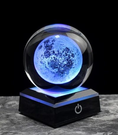 Lâmpada de cristal com efeito lunar azul, base preta com botão de ligar, iluminação suave e detalhes da superfície lunar visíveis.