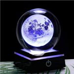 Bola de cristal com efeito lunar, iluminada com luz azul, sobre base preta com botão de ligar, fundo escuro.