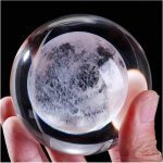 Bola de cristal transparente com efeito de superfície lunar, mostrando detalhes de crateras e manchas, segurada por uma mão com fundo escuro.