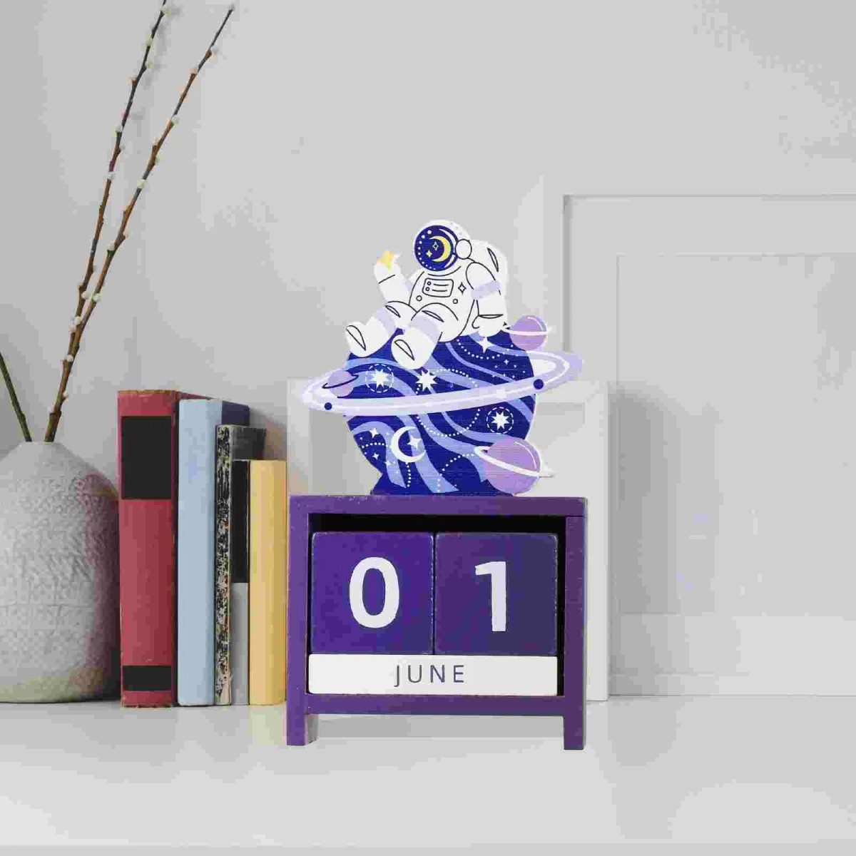 Calendário perpétuo com astronauta em estilo cartoon, decorativo, em tons de violeta e branco, com planetas e estrelas ao redor, em uma mesa branca ao lado de livros e uma planta.