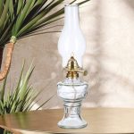 Candeeiro a óleo vintage com vidro transparente e chama artificial, colocado numa mesa de madeira com plantas verdes ao fundo.