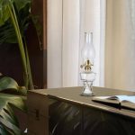 Candeeiro a óleo vintage de vidro transparente com chama artificial, colocado numa mesa de madeira ao lado de um livro aberto, com cortinas brancas ao fundo.