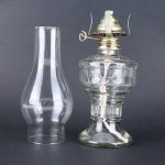 Lâmpada a óleo vintage de vidro transparente com componentes metálicos na parte superior, sem chama acesa, sobre fundo cinza escuro.