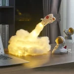 Foguete de brinquedo com luzes de plasma em forma de astronauta, emitindo luz amarela, colocado numa mesa de escritório com outros brinquedos espaciais ao fundo.