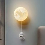 Lâmpada de parede com forma de lua cheia, com um pequeno astronauta pendurado por uma corda, decorando um quarto.