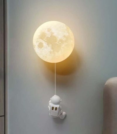 Lâmpada de parede com forma de lua cheia, com um pequeno astronauta pendurado por uma corda, decorando um quarto.