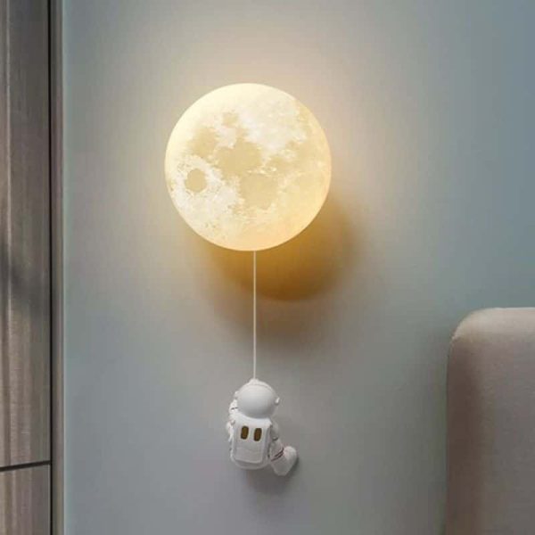 Lâmpada de parede com forma de lua cheia, com um pequeno astronauta pendurado por uma corda, decorando um quarto.
